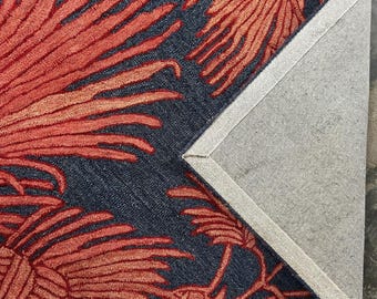 May include: A close-up of a rug with a navy blue background and a repeating pattern of red and orange palm leaves. The corner of the rug is folded back, revealing the gray backing.