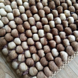 Op de afbeelding: Een handgemaakt tapijt met een getextureerd oppervlak, met een patroon van kleine, afgeronde, vervilte wollen ballen. Het kleurenpalet omvat tinten bruin, beige en crème, wat een warme uitstraling creëert. Het tapijt heeft een geweven rand.