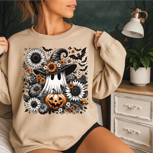 Peut inclure: Sweat-shirt beige avec un motif d'Halloween. Le motif représente un fantôme portant un chapeau de sorcière, entouré de tournesols, de citrouilles et de chauves-souris. Le motif utilise des couleurs noir, blanc, orange et jaune.
