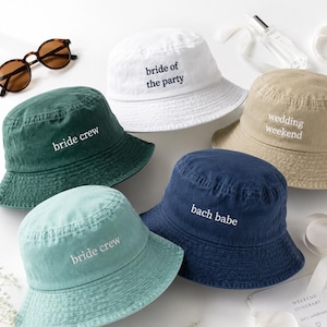 May include: Five bucket hats in white, dark green, light green, navy blue, and tan. The white hat says bride of the party, the dark green and light green hats say bride crew, the navy blue hat says bach babe, and the tan hat says wedding weekend.