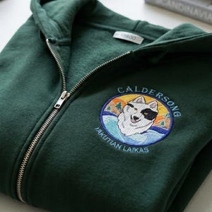 May include: A dark green zip-up hoodie with a detailed embroidered patch featuring a dog and the text "CALDERSONG YAKUTIAN LAIKAS". The hoodie is folded neatly, with a silver zipper. A dog bowl, leash, and books are in the background.