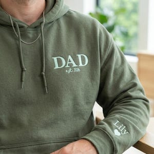 以下が含まれることがあります： 胸元にライトグリーンの文字で「DAD est. 2026」とプリントされたオリーブグリーンのパーカー。左袖にはライトグリーンの文字で「Lily」という名前が記されており、その下には小さなライトグリーンのハートのアイコンが添えられています。