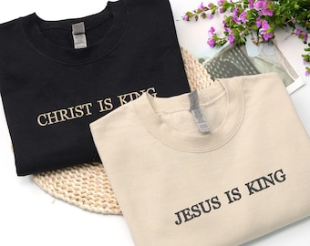 May include: Two crewneck sweatshirts, one black and one beige. The black sweatshirt has "CHRIST IS KING" embroidered in a light color, and the beige sweatshirt has "JESUS IS KING" embroidered in black. Both are displayed on a woven mat.