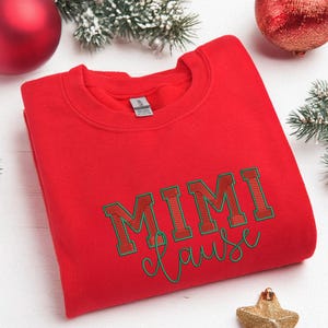 Puede incluir: Una sudadera roja con el texto "MIMI Clause" en letras verdes y rojas. La sudadera se muestra sobre una superficie blanca con adornos navideños, incluyendo bolas y ramas.