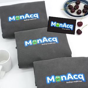 May include: Three folded gray sweatshirts with the embroidered text "MonAcq duolingo english test" in blue and white. A small graphic of a green owl is in the center of the text. A small black square with the same logo is also present. Cherries are on a plate.