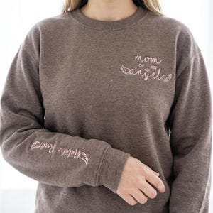 A woman wearing the Dark Heather Gildan sweatshirt, showcasing the pink "Mom of an Angel" chest embroidery and the personalized sleeve. The heathered fabric provides a soft, comfortable texture. This image highlights the sweatshirt as a wearable memorial, perfect for those looking for aesthetic miscarriage awareness apparel and comforting grief gifts.