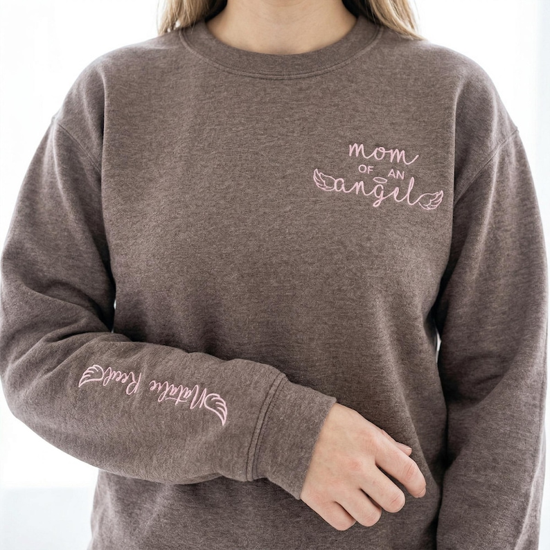 A woman wearing the Dark Heather Gildan sweatshirt, showcasing the pink "Mom of an Angel" chest embroidery and the personalized sleeve. The heathered fabric provides a soft, comfortable texture. This image highlights the sweatshirt as a wearable memorial, perfect for those looking for aesthetic miscarriage awareness apparel and comforting grief gifts.