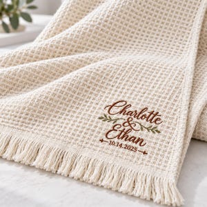 May include: A cream-colored waffle-weave blanket featuring fringe trim and brown embroidery that reads Charlotte & Ethan with the date 10.14.2025, accented by small green leaf sprigs positioned between the names and on the sides.