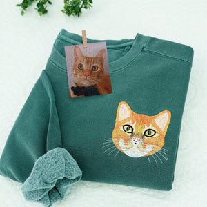 Puede incluir: Sudadera verde azulado con un bordado de la cara de un gato atigrado naranja en la parte delantera. Una pequeña foto de un gato similar está sujeta a la sudadera. La manga de la sudadera está doblada.