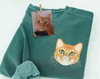 Puede incluir: Sudadera verde azulado con un bordado de la cara de un gato atigrado naranja en la parte delantera. Una pequeña foto de un gato similar está sujeta a la sudadera. La manga de la sudadera está doblada.