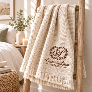 May include: A cream-coloured waffle-knit throw blanket with fringe edges, featuring brown embroidery of a laurel wreath surrounding the monogram EL, the names Emma & Liam, and the date 09.08.2023, draped over a wooden ladder.