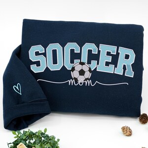 Navy blue Soccer Mom sweatshirt with varsity light blue letters and soccer ball graphic, sleeve embroidered with simple heart icon. Cozy personalized gift for soccer moms, perfect for chilly match days and supportive parents in youth soccer teams.