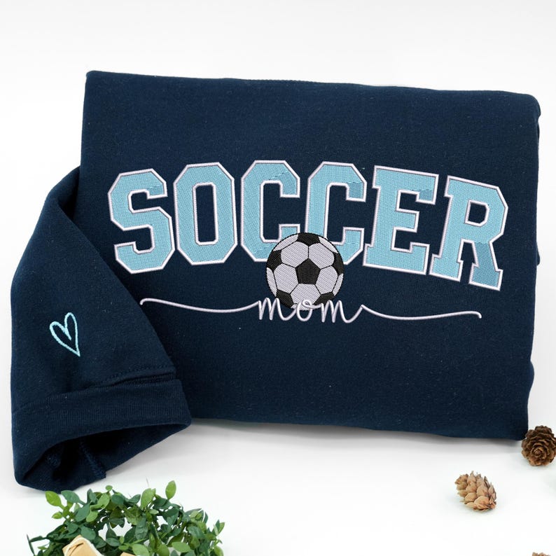 Navy blue Soccer Mom sweatshirt with varsity light blue letters and soccer ball graphic, sleeve embroidered with simple heart icon. Cozy personalized gift for soccer moms, perfect for chilly match days and supportive parents in youth soccer teams.