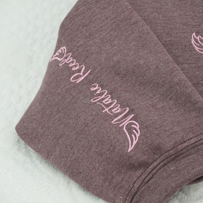 Detailed shot of the custom sleeve embroidery on a heathered brown sweatshirt. The name "Natalie Reed" is stitched in light pink thread, framed by elegant angel wings. This "name on sleeve" personalization is a popular trend for custom memorial clothing, offering a subtle and meaningful way to keep a loved one's memory close to the heart.