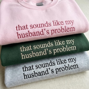 May include: Three sweatshirts stacked on top of each other. The top sweatshirt is light pink, the middle one is dark green, and the bottom one is gray. Each sweatshirt has the embroidered text "that sounds like my husband's problem".