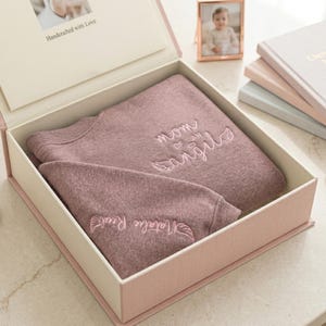 A personalized "Mom of an Angel" sweatshirt in Dark Heather, neatly folded inside a high-end cream gift box. The packaging includes a "Handcrafted with Love" insert, making it a perfect ready-to-gift option for infant loss support or memorial services. High-intent keywords: custom remembrance gift, miscarriage gift for mom, and memorial packaging.