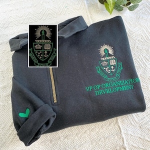May include: Black sweatshirt with a green embroidered crest and the text "VP OF ORGANIZATION DEVELOPMENT." The sweatshirt has a gold zip and a green heart detail on the cuff. The crest includes a candle, book, and anchor.