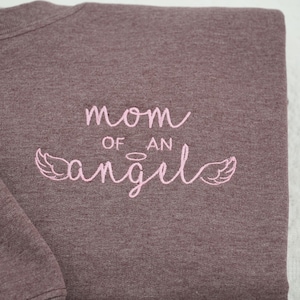 Close-up of high-quality pink embroidery on a Dark Heather Gildan 18000 crewneck. The design reads "mom of an angel" in a delicate script font, accented with a small halo and angel wings. This custom embroidered memorial apparel is a top choice for those searching for pregnancy loss keepsakes or sentimental bereavement gifts for mothers.