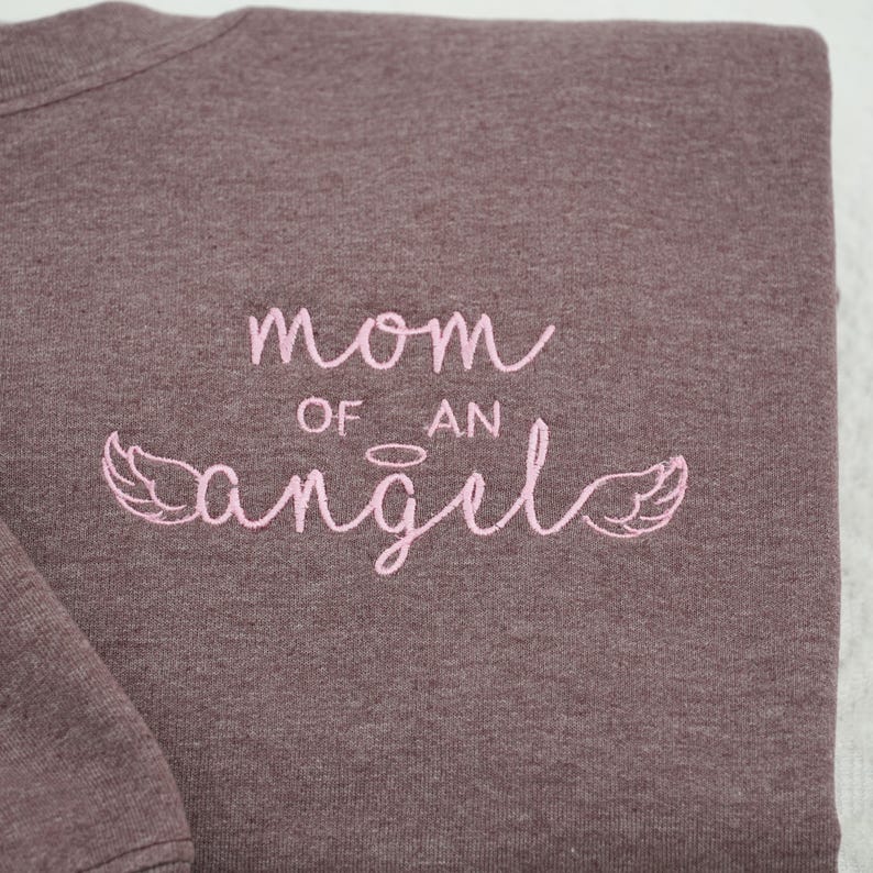 Close-up of high-quality pink embroidery on a Dark Heather Gildan 18000 crewneck. The design reads "mom of an angel" in a delicate script font, accented with a small halo and angel wings. This custom embroidered memorial apparel is a top choice for those searching for pregnancy loss keepsakes or sentimental bereavement gifts for mothers.