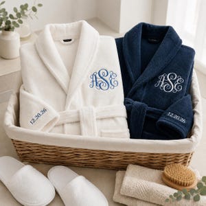 May include: A white bathrobe with blue monogram embroidery and a navy blue bathrobe with white monogram embroidery, both featuring the date 12.20.26 on the sleeve, placed in a woven basket with white slippers and folded towels nearby.
