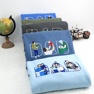 Stack of embroidered “DAD” sweatshirts in blue, green, grey, and light blue. Each letter “DAD” is filled with custom father-and-child illustrations. Personalized Father’s Day or birthday gift, cozy cotton crewnecks for dads, sentimental keepsake apparel.