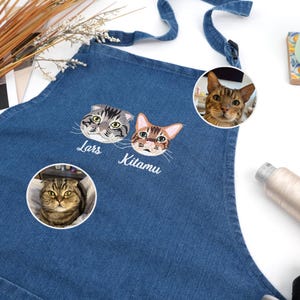 May include: Blue denim apron featuring embroidered cat faces with the names "Lars" and "Kitamu" below. Additional cat portraits are displayed in circular frames. The apron has a neck strap and waist ties.
