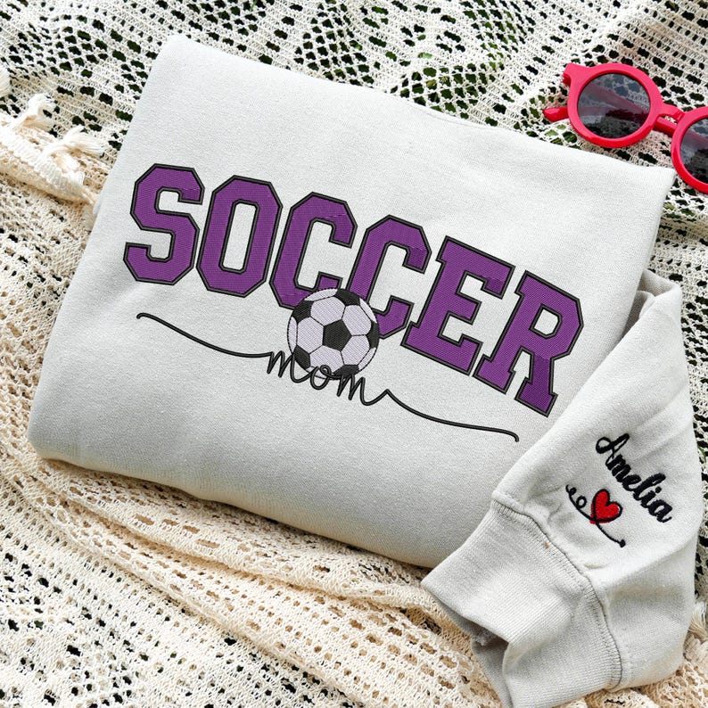Sand Soccer Mom sweatshirt with purple varsity lettering and soccer ball design, personalized with “Amelia” embroidered on the sleeve and red heart accent. Styled on lace fabric background with red sunglasses, making a thoughtful gift for proud soccer moms and sports moms.