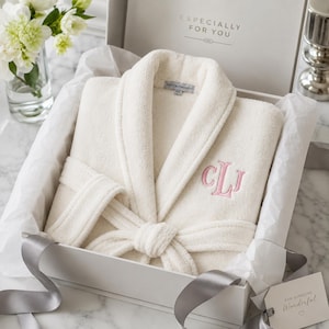 May include: A cream-colored plush robe with a shawl collar and a matching tie belt, featuring a pink embroidered monogram of the letters CLJ on the chest. The robe is presented inside a white gift box with the text Especially For You.
