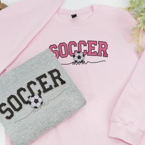 Light pink Soccer Mom sweatshirt with bold varsity-style pink letters and soccer ball graphic, personalized with embroidered sleeve text “Miss Isabell.” Folded gray option included, showing black lettering with soccer ball. Perfect custom gift for soccer moms supporting kids’ games.