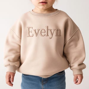 Puede incluir: Sudadera beige con el nombre "Evelyn" bordado en un tono a juego. La sudadera tiene cuello redondo y mangas largas. El niño lleva vaqueros azules.