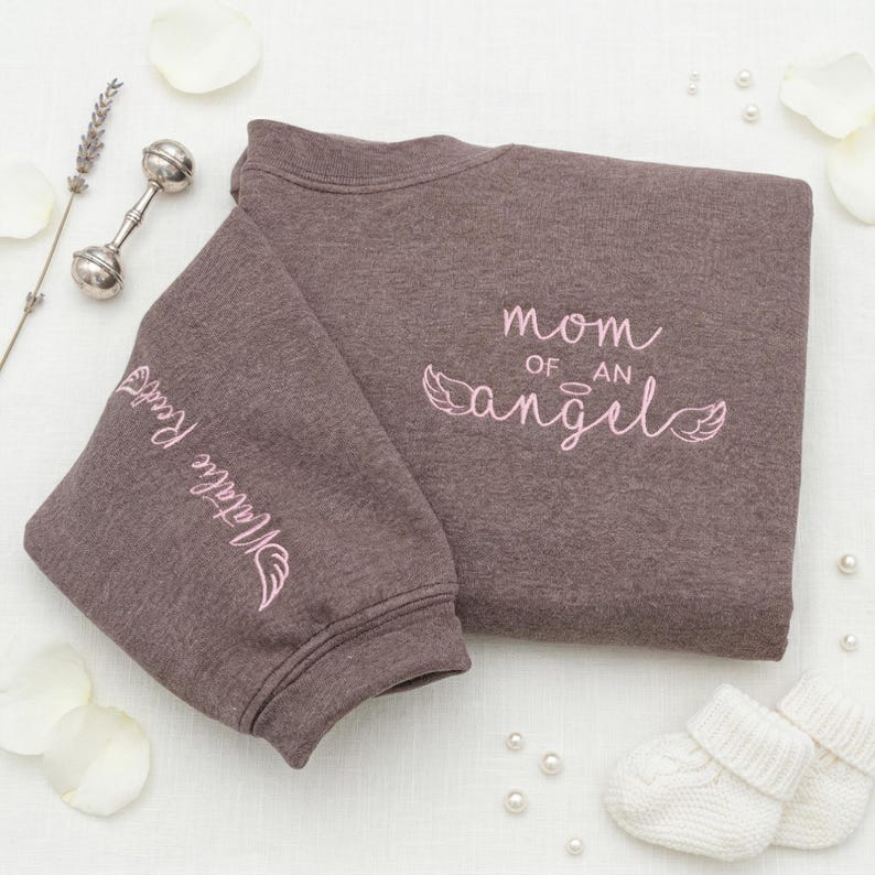 A Dark Heather Gildan sweatshirt featuring pink embroidery with the text "Mom of an Angel" and decorative wings. The sleeve is personalized with the name "Natalie Reed." This personalized angel mom sweatshirt serves as a beautiful memorial gift for miscarriage or infant loss, styled with white baby booties and petals for a gentle, aesthetic look.