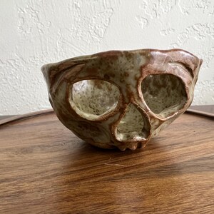 May include: A handmade ceramic bowl in the shape of a skull. The bowl has a textured surface with brown and tan colors. The eye sockets and nose cavity are open, creating a unique design. The bowl is placed on a wooden tray.