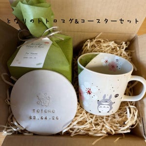 May include: A gift set featuring a ceramic mug and coaster with a Totoro theme. The mug has a white background with cherry blossom and Totoro illustrations. The coaster is white with the word "TOTORO" and a date. The set is packaged in a cardboard box.