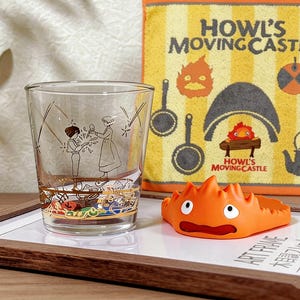Puede incluir: Un vaso transparente con detalles dorados y una ilustración de dibujos animados. Un personaje de dibujos animados naranja y una toalla amarilla y naranja con el texto "Howl's Moving Castle" también son visibles. Los artículos están sobre una bandeja de madera.