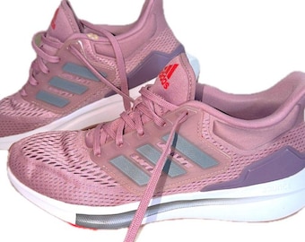 May include: A pair of mauve athletic sneakers featuring a breathable mesh upper, three grey side stripes, and a white midsole with the word Bounce embossed on the heel. The tongue displays a red Adidas logo with matching laces.