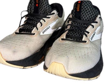 May include: A pair of Brooks Ghost 16 running shoes featuring a light beige mesh upper, black laces, and a black midsole. The tongue displays the text GHOST 16 in orange, and the side of the shoe features the Brooks logo in white.