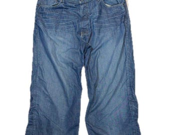 May include: Blue denim capri pants featuring a mid-rise waist, a button and zipper fly closure, and five pockets. The fabric displays a faded wash with horizontal whiskering patterns across the upper thighs and straight-cut leg openings.