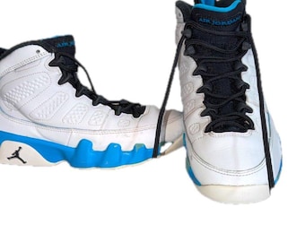 May include: A pair of high-top athletic sneakers featuring a white leather upper with a bright blue midsole and black laces. The tongue displays the text Air Jordan in blue, and a black Jumpman logo is visible on the side of the heel.