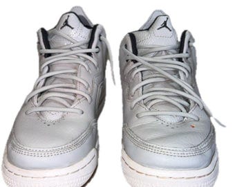 May include: A pair of light gray leather sneakers featuring a high-top silhouette, matching gray laces, and a black Jumpman logo on the tongue. The shoes have a textured leather upper, a white rubber midsole, and a perforated toe box design.