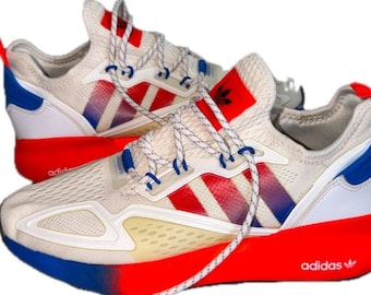 May include: A pair of off-white sneakers featuring red and blue gradient stripes on the sides, white laces, and a bright red midsole. The heel area includes blue accents, and the Adidas logo is printed on the tongue and the side of the sole.