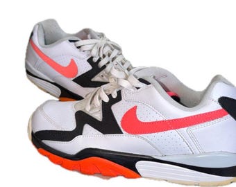 May include: A pair of white athletic sneakers featuring black overlays, a bright orange Nike swoosh logo on the side, white laces, and a multi-colored sole with orange and black sections. The shoes have a perforated side panel and a low-top design.