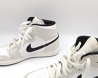 May include: A pair of high-top sneakers featuring a white leather base with light gray textured overlays. The shoes include black Nike swoosh logos on the sides, black interior lining, and white laces. The design is finished with a white midsole and a black outsole.