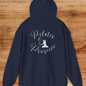 Puede incluir: Sudadera con capucha azul marino. La parte trasera presenta el texto blanco "Pilates & Prosecco" en fuente cursiva, con una pequeña ilustración de una persona haciendo pilates. La sudadera está hecha de un material suave.