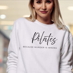 May include: A white long-sleeve crewneck sweatshirt featuring black text on the front. The word Pilates is written in a large, cursive script font, while the phrase BECAUSE MURDER IS WRONG is printed below in a smaller, clean sans-serif font.