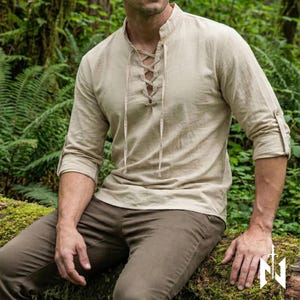 May include: Beige long-sleeved shirt with a lace-up neckline and rolled-up sleeves. The shirt is made of a light, textured fabric and is paired with brown trousers. The style is reminiscent of historical or medieval attire.