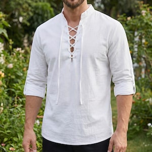 Pode incluir: Uma camisa de linho branca com gola mandarim e atacadores na frente. As mangas compridas est&atilde;o enroladas. A camisa &eacute; feita de um tecido leve.