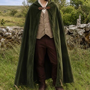 May include: A floor-length dark olive green hooded cloak with a brown leather clasp at the neck, worn over a beige button-up vest and dark brown trousers. The cloak features a smooth texture and drapes down to the ankles, providing full coverage.