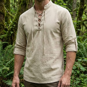 Pode incluir: Camisa bege de manga comprida com decote com atacadores. A camisa tem gola mandarim e as mangas est&atilde;o enroladas. A camisa &eacute; feita de um tecido leve, adequado para climas quentes. O fundo &eacute; uma exuberante floresta verde.