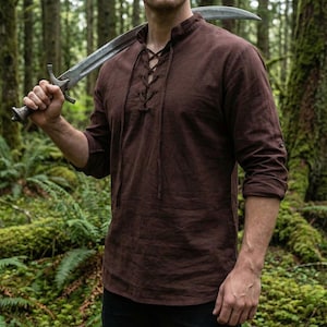 Pode incluir: Uma camisa marrom de manga comprida com decote com cord&otilde;es e gola mandarim. As mangas est&atilde;o enroladas. A imagem mostra uma camisa de estilo medieval, adequada para cosplay ou reconstitui&ccedil;&otilde;es hist&oacute;ricas.