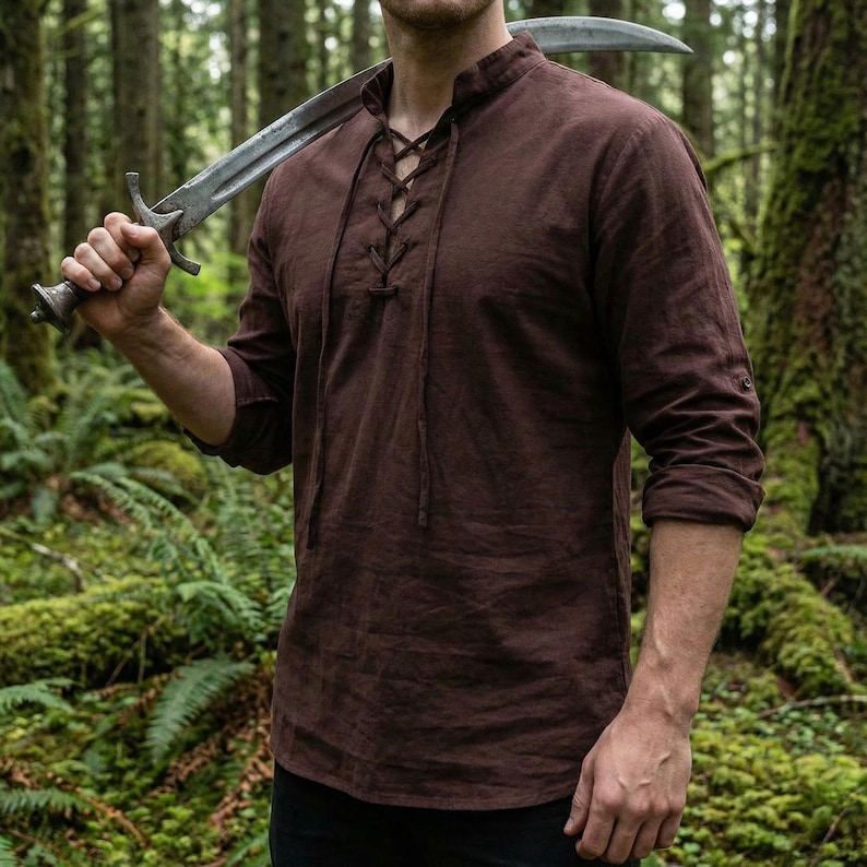 Pode incluir: Uma camisa marrom de manga comprida com decote com cord&otilde;es e gola mandarim. As mangas est&atilde;o enroladas. A imagem mostra uma camisa de estilo medieval, adequada para cosplay ou reconstitui&ccedil;&otilde;es hist&oacute;ricas.